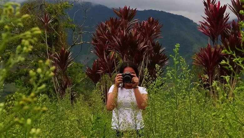 A picture of a woman in a white t-shirt standing in a lush green tropical field taking a picture  of the photographer