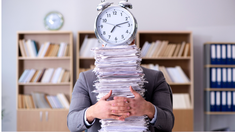 A person hides behind a stack of papers with a clock balanced on top.