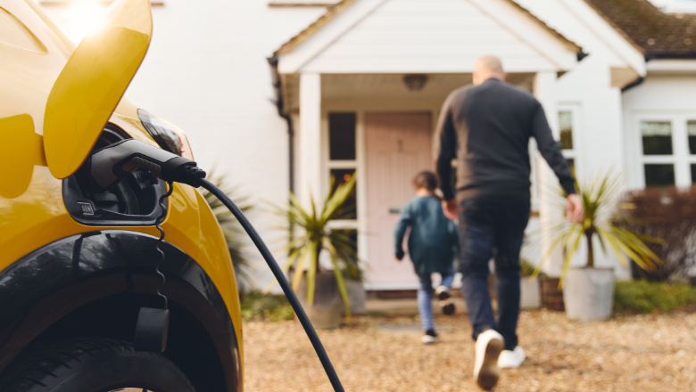 A yellow electric vehicle is charging on a driveway whilst a father and son walk into their house.