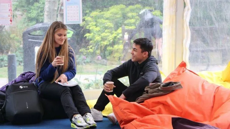 Two people sitting on bean bags in a relaxed indoor setting, both holding paper cups and smiling at each other.