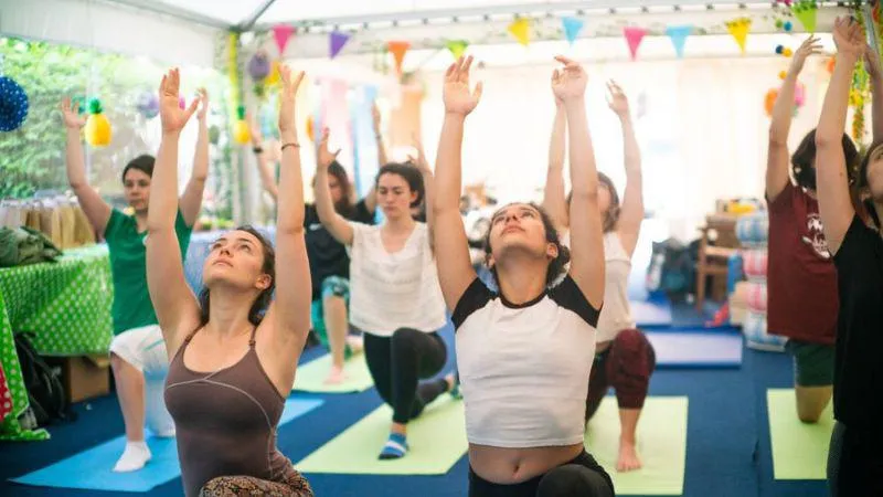 A group yoga class in progress, with participants in an indoor space raising their arms overhead as they stretch. The room is decorated with colorful flags and bright lights, creating a cheerful atmosphere.