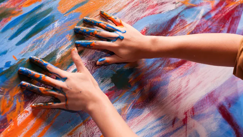 Artist's hands covered in paint against the abstract-coloured canvas.