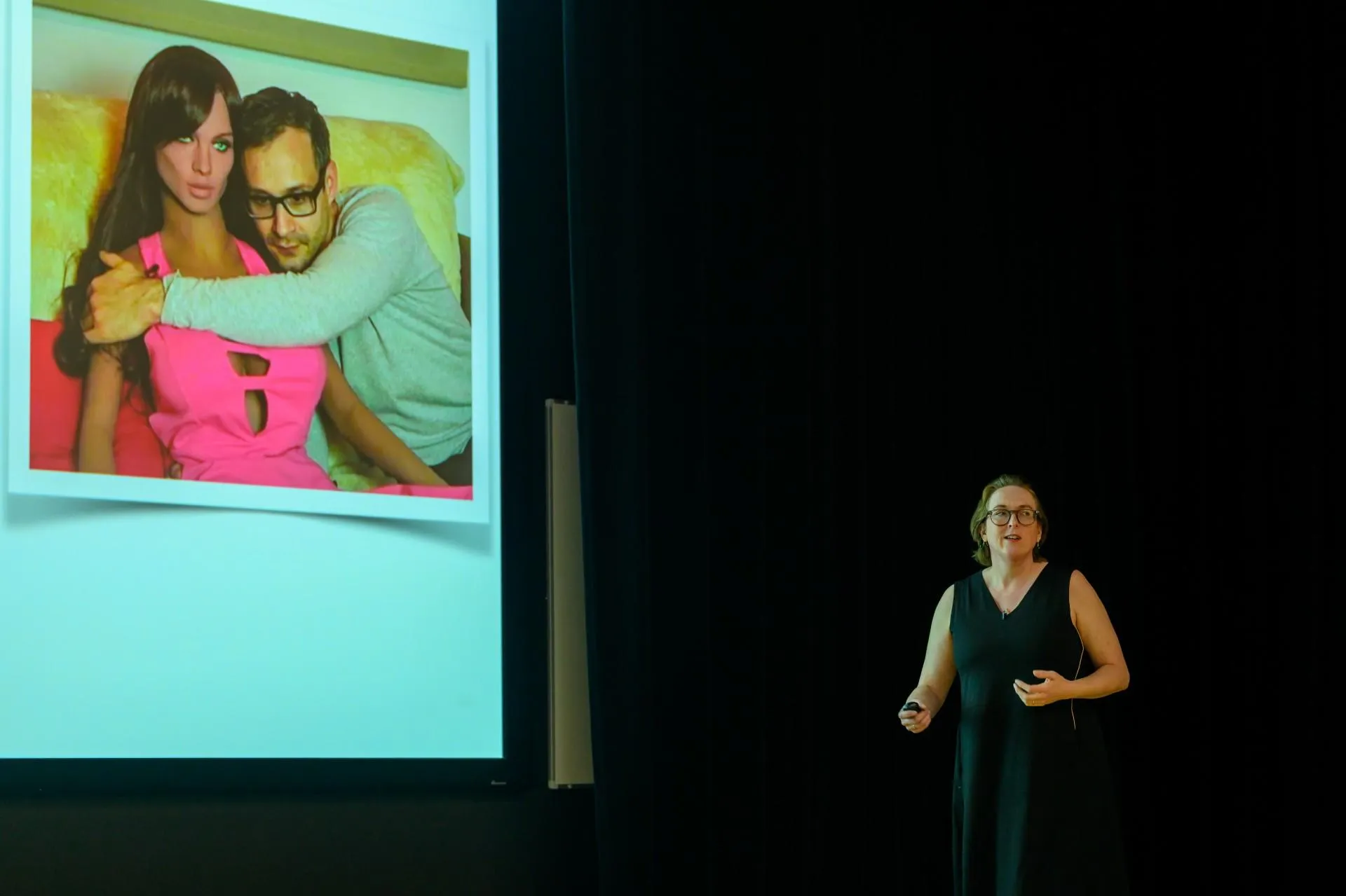 A white woman in a black sleeveless dress is presenting to an audience. She is stood to the right of a projected slide, which shows a human man holding a sex doll in the style of a woman.