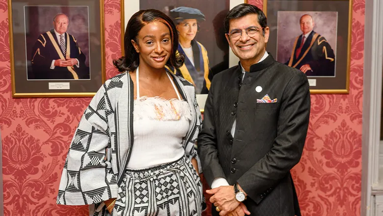 Cuppy, a woman with dark hair, wearing black and white clothing, stands next to Shitij Kapur, a man with neat, dark hair. Both are smiling in front of several portraits.