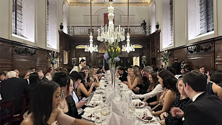 A long dining table surrounded by elegantly dressed guest at a formal event in a grand hall.