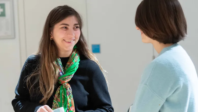 A brunette woman with long hair, in a black sweater, and a green scarf, smiling while talking to the person next to them