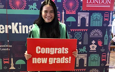 A woman with long black hair smiles holding a red placard that reads congrats new grads