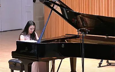 A woman with long black hair plays a grand piano on stage