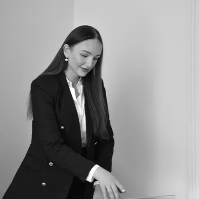 A confident-looking young woman in a relaxed business suit sits in an office at a desk with a computer in front of her