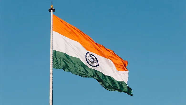 The flag of India flies on a white flagpole in front of a brilliant blue sky.