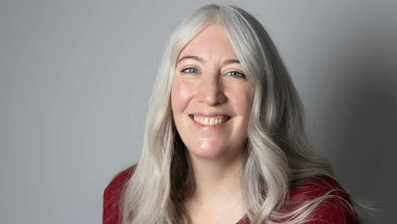 A woman with long grey hair, wearing a red top, smiles warmly