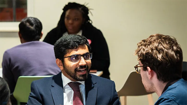 A man with dark hair and glasses, wearing a sharp red tie and suit jacket, speaks to another attendee at a mentoring event