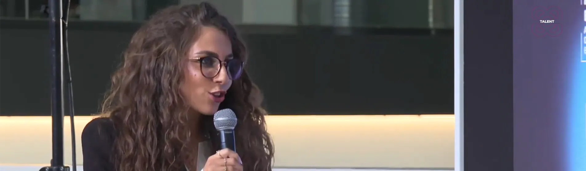 A young woman with long, dark hair and wearing a dark blue jacket stands on a stage at a presentation and talks to the audience. She holds a microphone in her right hand