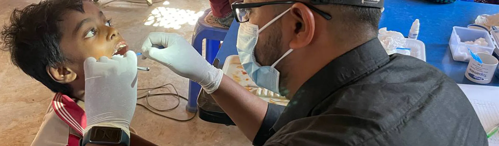 A man wearing a face mask and surgical gloves uses dental instruments to examine the mouth of a child in a refugee camp