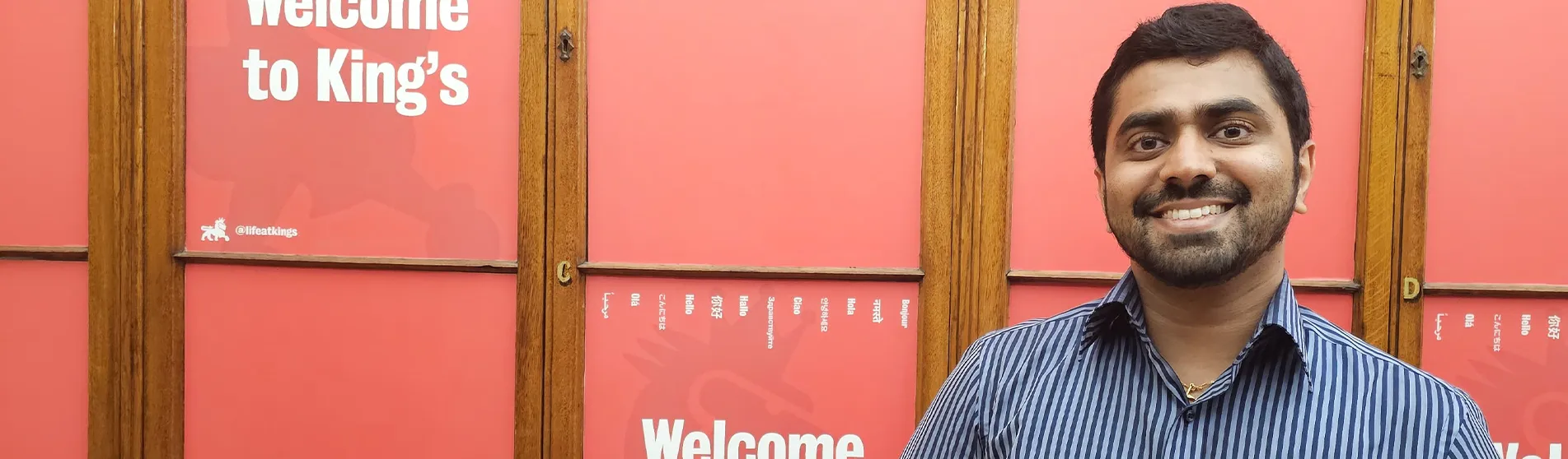 A smiling young man wearing a short-sleeved shirt stands in front of a red wall with Welcome to King’s written on it