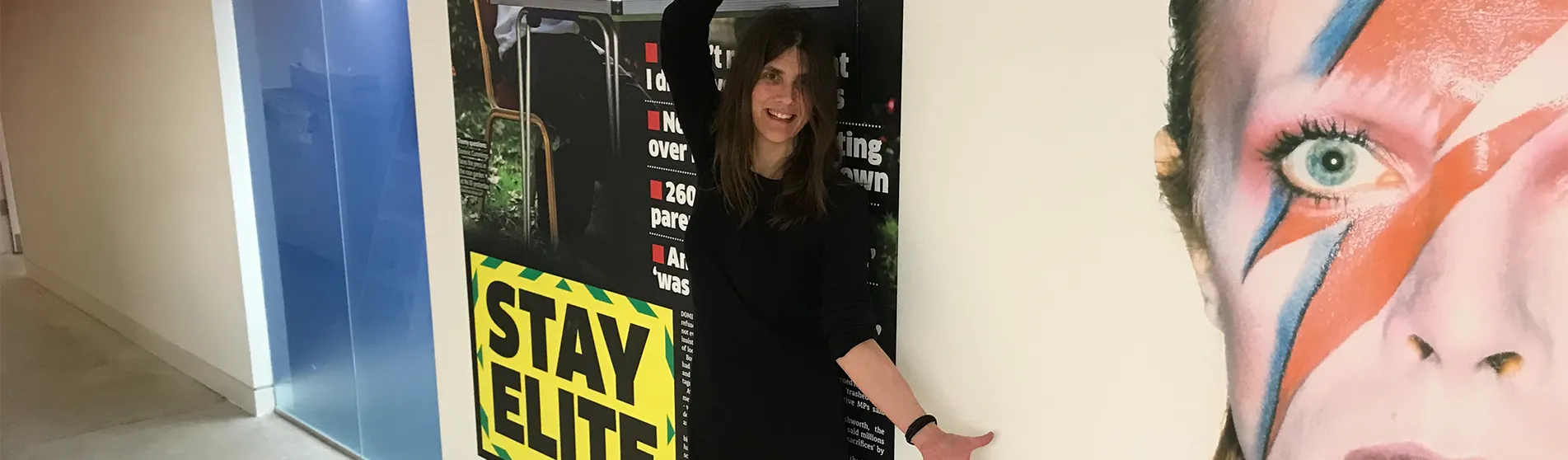 A young woman with long hair and a stylish black dress smiles as she stands in front of two huge framed posters of different front pages from Metro newspaper