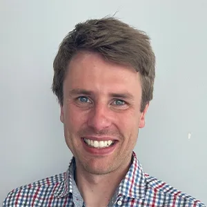 A man smiles widely, wearing a checked shirt of white, blue and red