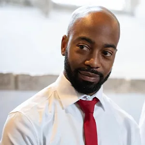 A man with a dark beard looks to his side. His is wearing a white shirt and red tie.