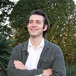 A man wearing a dark green jacket and white shirt, crosses his arms and smiles as he stands in front of lush green bushes