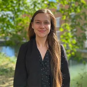 Standing in front of a vibrant background of green leaves and grass, a woman with long brown hair, wearing a dark polka dot shirt under a black jacket, smiles