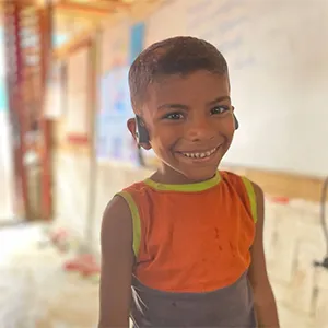 A child wearing a t-shirt has a hearing device worn over his ears and smiles at the camera