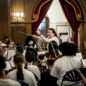 Lucy, wearing a white shirt, directs an orchestra, with string instruments in the foreground