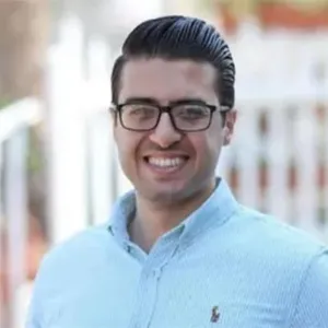 A man with slicked back black hair, wearing dark glasses and a light blue shirt, smiles