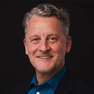 A man with cropped grey hair, wearing a deep blue shirt under a black jacket, smiles at the camera