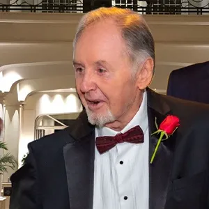 An older man in a black tie suit, wearing a maroon bowtie and a rose on his lapel