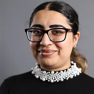 A woman with black glasses and wearing a white beaded necklace smiles at the camera