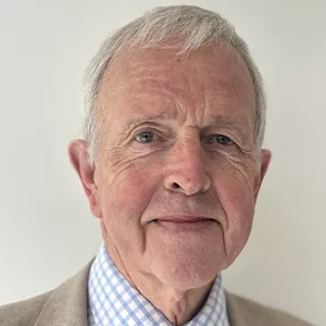 An older man wearing a blue and white checkered shirt under a beige jacket looks warmly at the camera