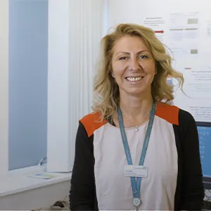 A woman with medium length blonde hair, wearing a white and orange shirt with black sleeves, and a light blue lanyard, stands in a doctor's office