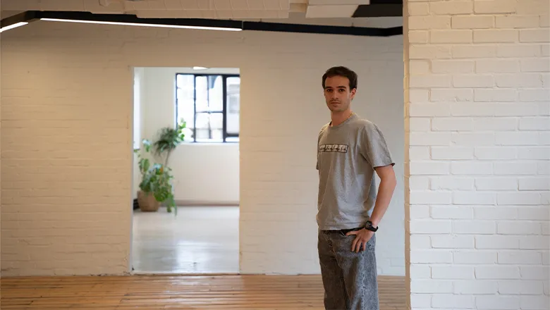 A young man with dark hair wearing smart and casual clothes stands in a clean and uncluttered office space.