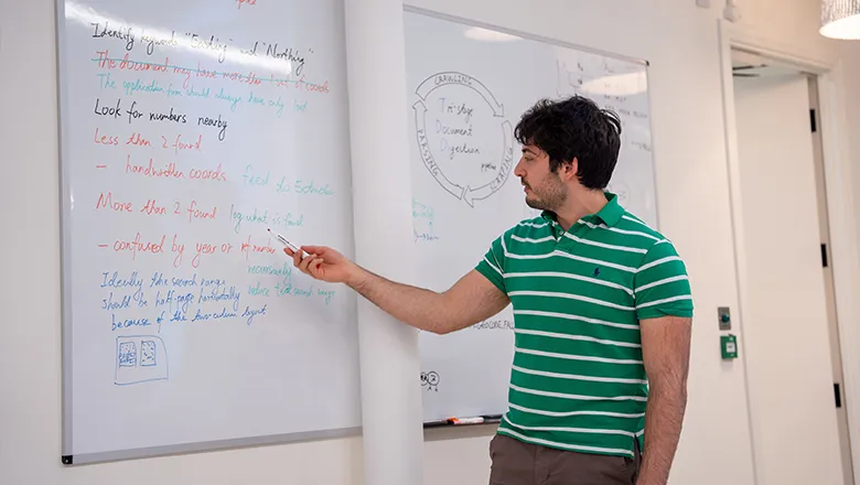 A young man wearing smart and casual clothes stands in front of a whiteboard adding comments with a marker pen to a plan.