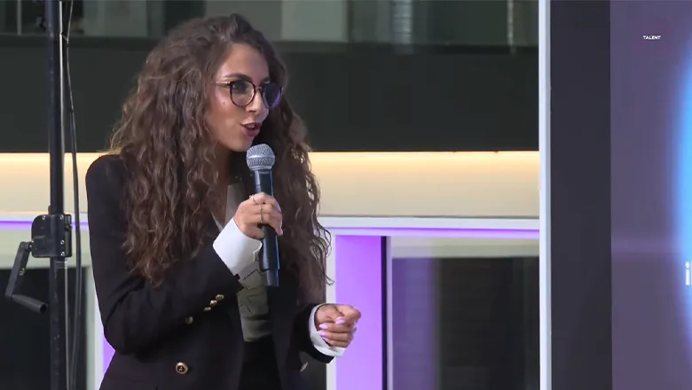 A young woman with long, dark hair and wearing a dark blue jacket stands on a stage at a presentation and talks to the audience. She holds a microphone in her right hand