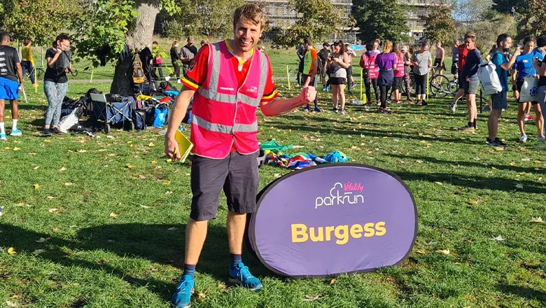 A man in a bright pink high visibility jacket, dark shorts and blue shoes, smiles next to a sign that says 'Burgess' in a park