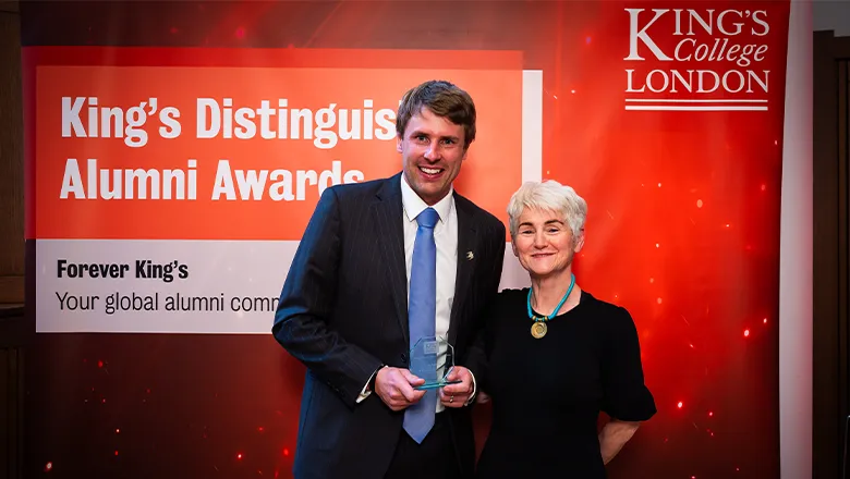 A man and a woman stand in front of a red background with the words King's Distinguished Alumni Awards on it. The man, wearing a smart jacket with light blue tie, smiles as he holds an award