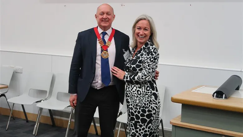 A man in a suit stands next to a woman in black and white dress. Both smile and he proudly displays a ceremonial chain of office that is hung around his neck