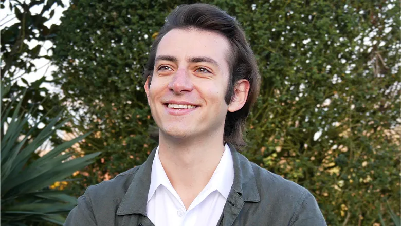 A man wearing a dark green jacket and white shirt, crosses his arms and smiles as he stands in front of lush green bushes