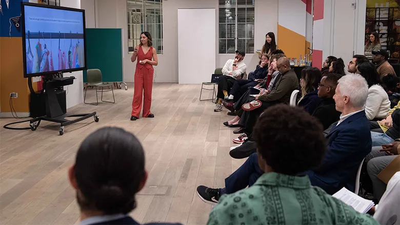 A young woman with dark hair and wearing matching orange trousers and a top makes a presentation in front of a packed group of people. On the left is a screen with a picture of clothes hanging from a rail.