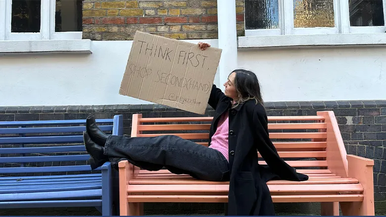 A young woman with dark hair sits on a bench at an outdoor location. She wears dark blue jeans, a pink jumper and a black coat. In her hands is a sign that reads:‘Think first, shop second (hand).’