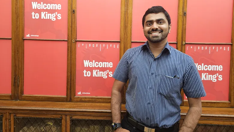 A smiling young man wearing a short-sleeved shirt stands in front of a red wall with Welcome to King’s written on it