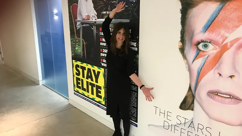 A young woman with long hair and a stylish black dress smiles as she stands in front of two huge framed posters of different front pages from Metro newspaper