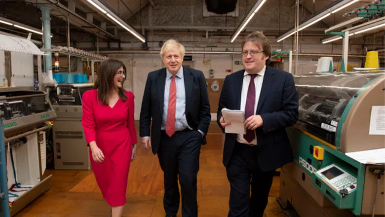 A young woman with long hair and a red dress walks alongside former Prime Minister Boris Johnson and another man. Both men wear suits.