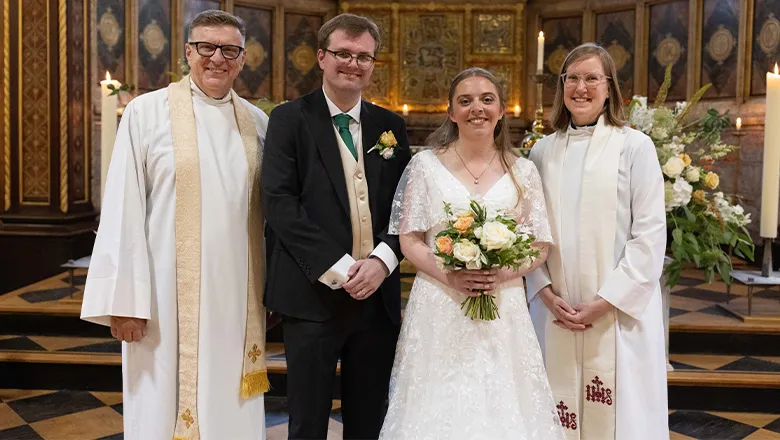 A couple, just married, stand holding flowers next to two members of the clergy