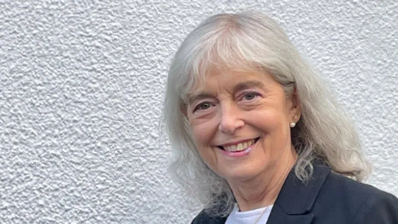 A woman with grey hair standing against a white wall smiles at the camera