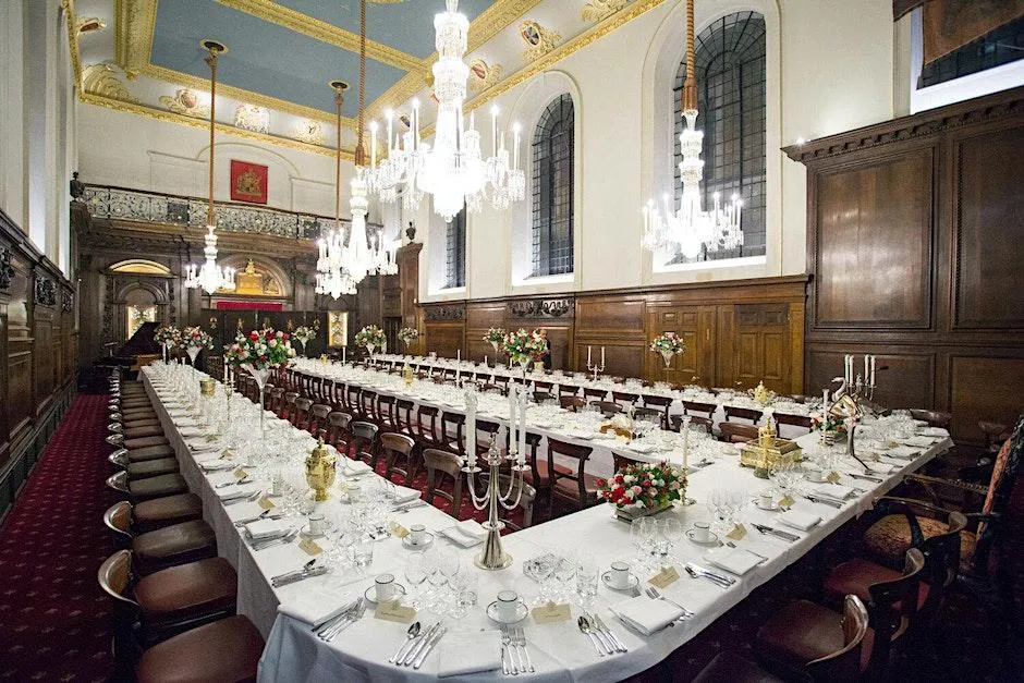 A three-pronged table is dressed for dinner, covered in a white cloth, in a large, old hall adorned with wood panelling.