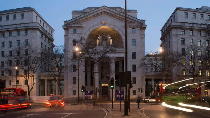 Bush House building with a central illuminated archway, flanked by symmetrical wings. Light trails from vehicles and buses add motion to the evening scene, framed by leafless trees under a twilight sky.