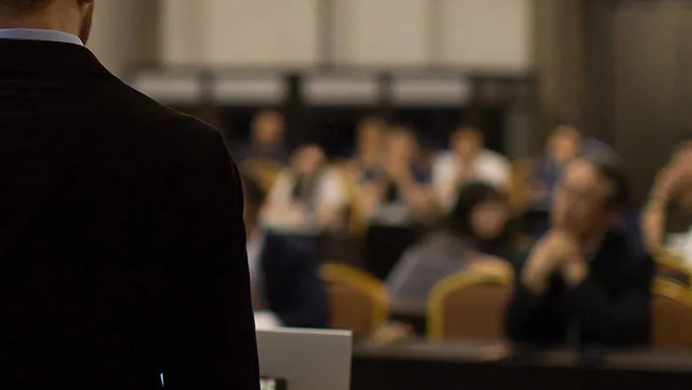 A man in a suit stands before a group of attentive people in a conference setting.