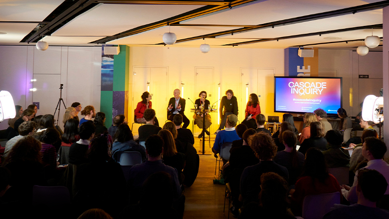 5 panellists in front of yellow-orange lighting and audience with a screen showing "Cascade Inquiry"